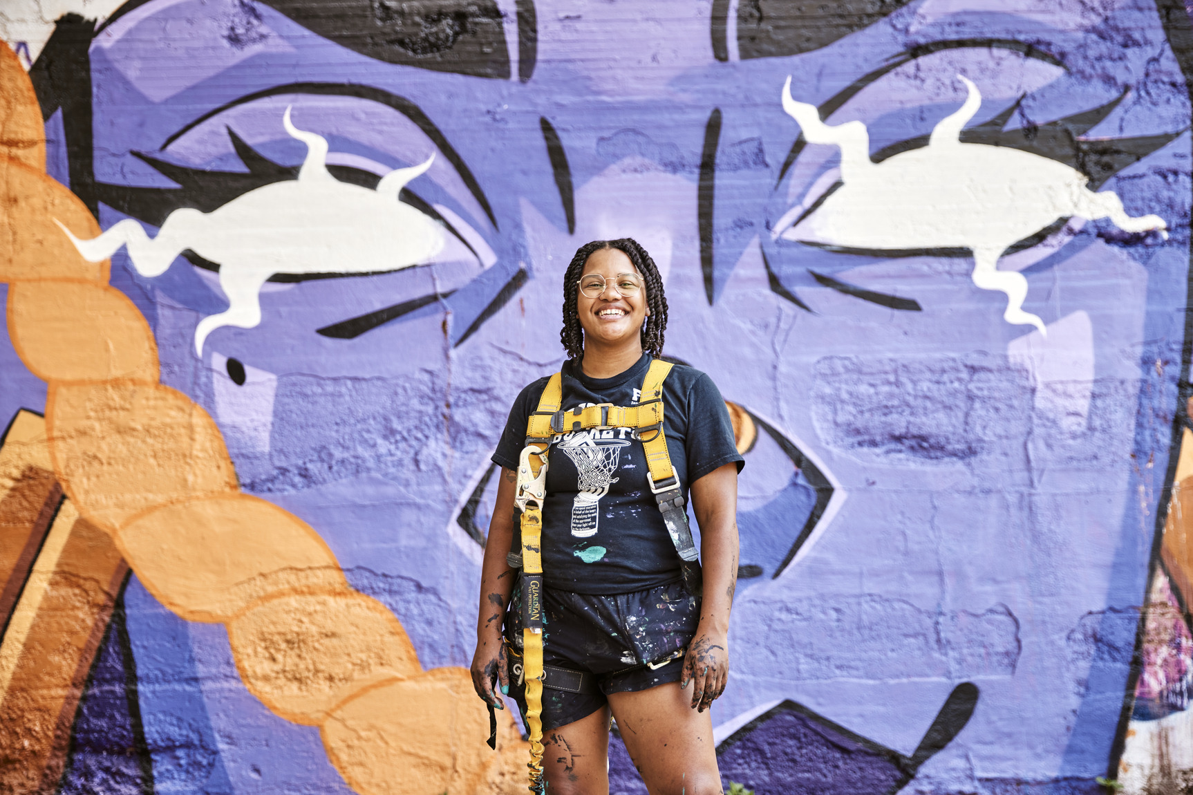 An artists stands, smiling and covered in paint, in front of a colorful mural.