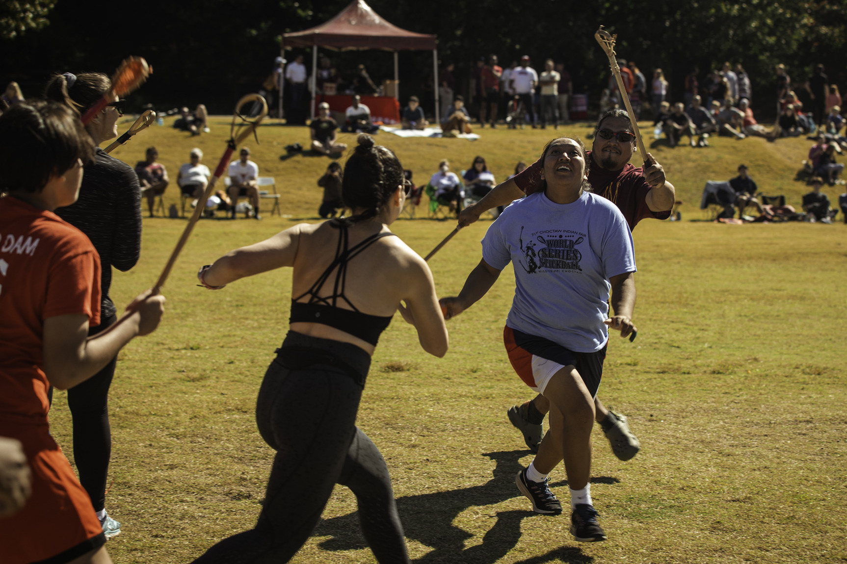 A group of people play Stickball on a field while spectators watch on.