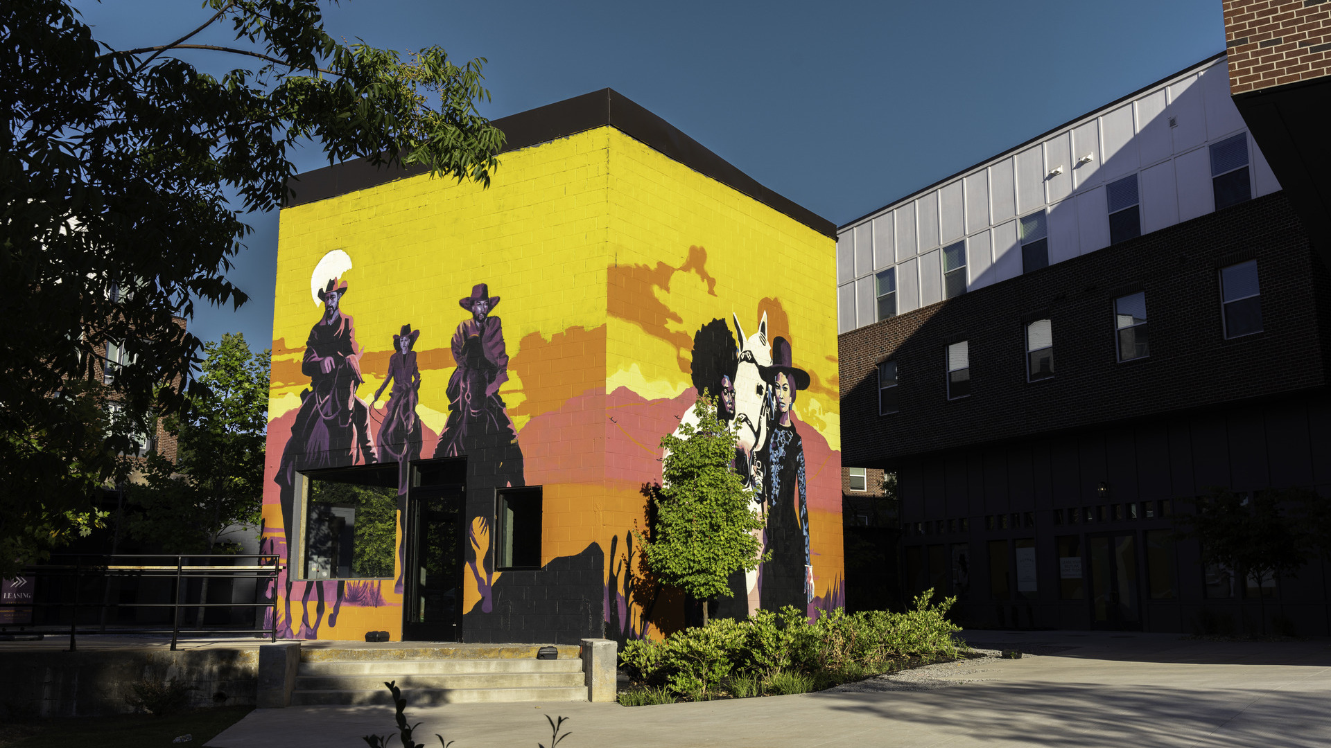 An image of two vibrantly colored murals on adjacent sides of a small building, surrounded by greenery and buildings. One mural features three figures on horseback wearing cowboy hats, the other showing two women standing with a horse, both set against a backdrop of mountains and sky.