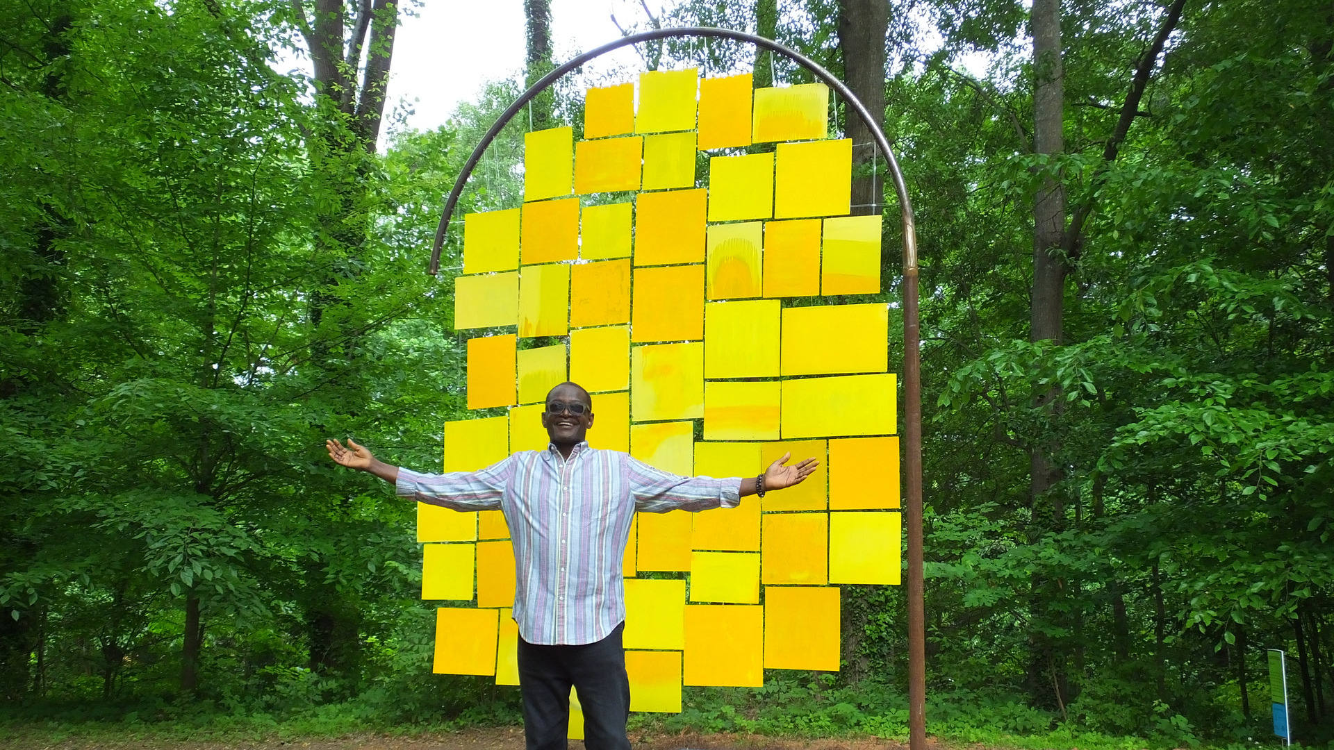 A man stands proudly in front of a sculpture containing yellow square and rectangular panels hanging from a rounded metal structure.