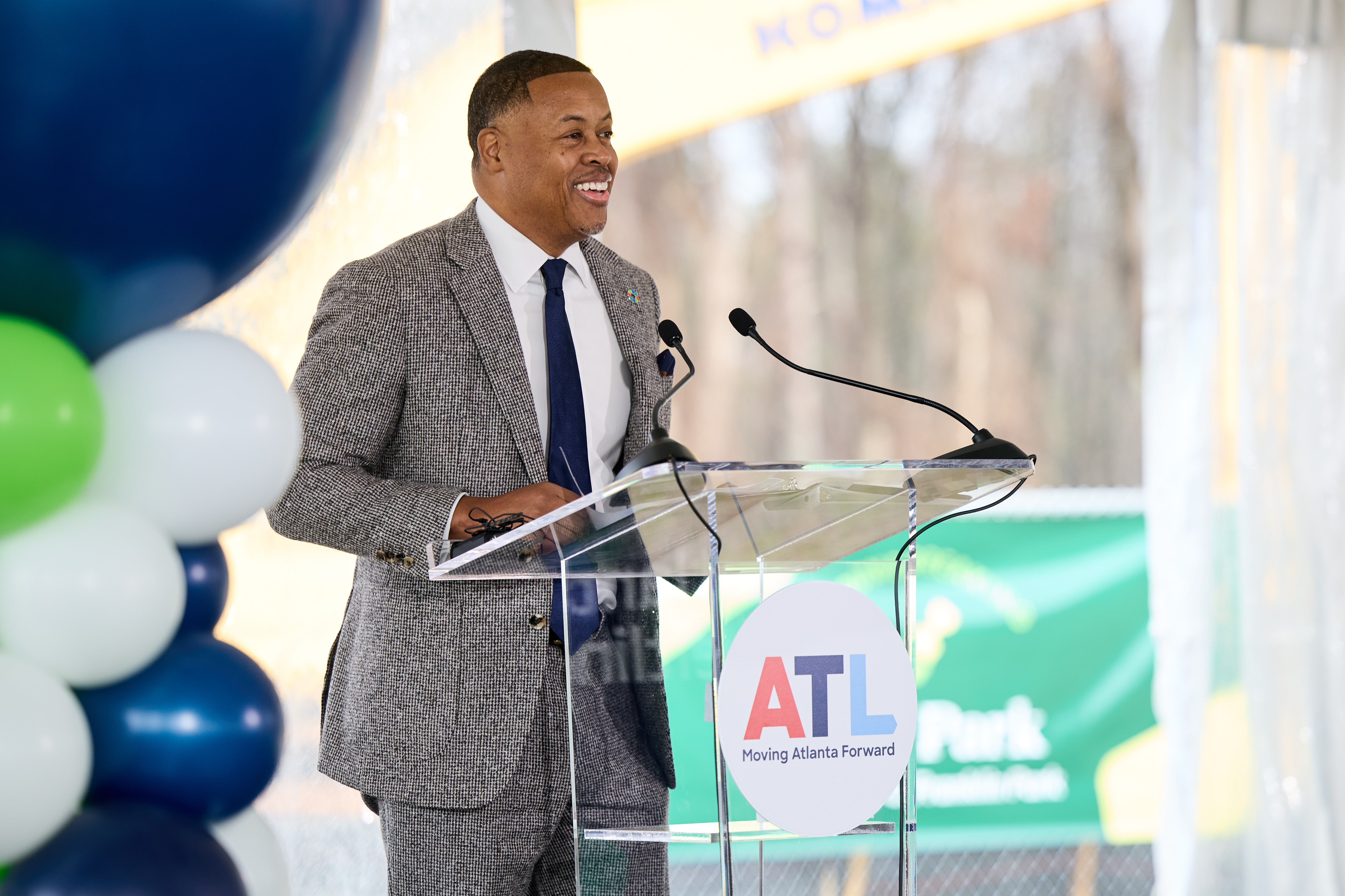 Atlanta President and CEO Clyde Higgs in a gray suit and blue tie speaks at a podium with an "ATL Moving Atlanta Forward" logo. Balloons and banners are in the background.
