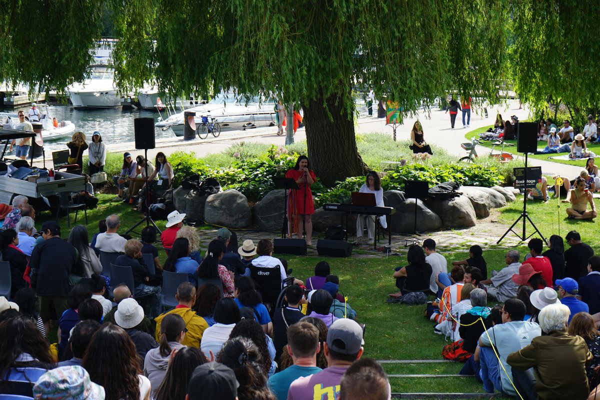 Outdoor concert with a woman singing and another playing keyboard under a large tree. A seated audience watches, with boats docked in the background.