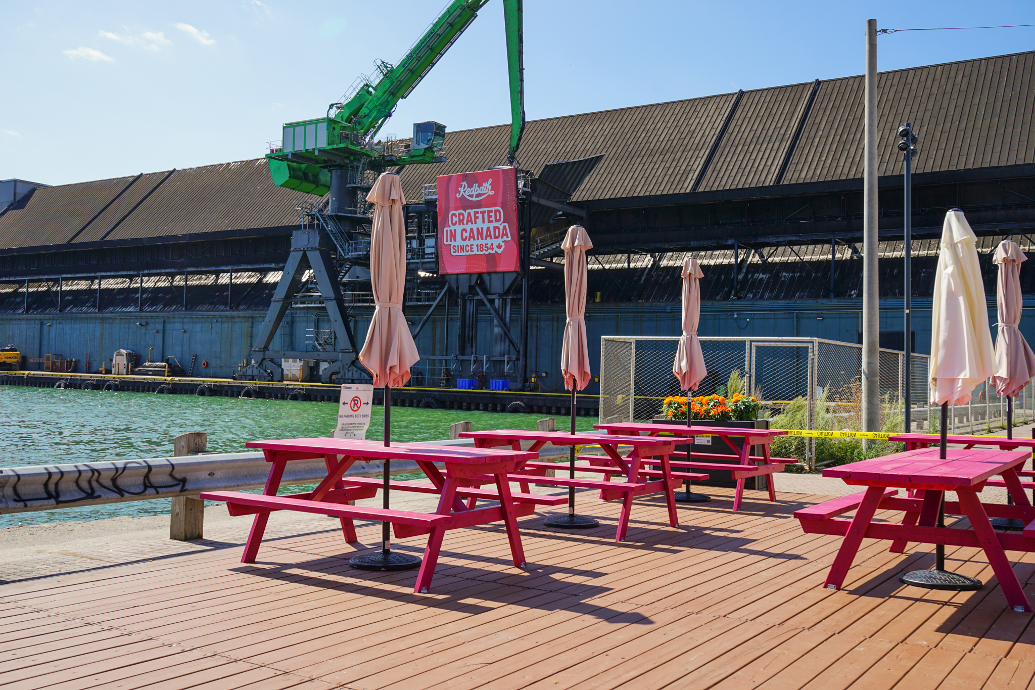 Red picnic tables with closed umbrellas on a wooden deck by the water, industrial building and crane in the background, with a "Crafted in Canada" sign.