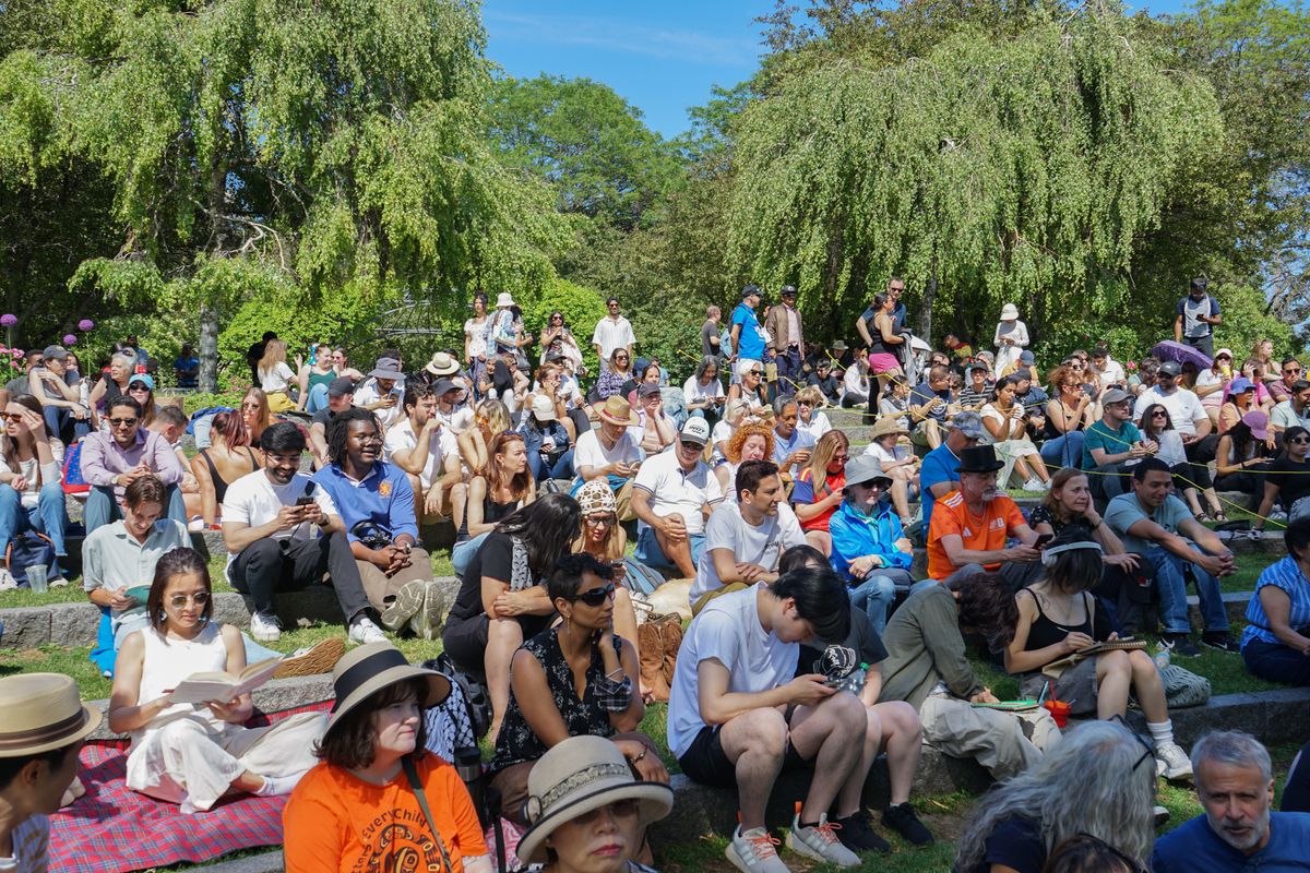 A diverse group of people sitting on grassy steps outdoors, some reading or using phones, under sunny skies and surrounded by leafy trees.