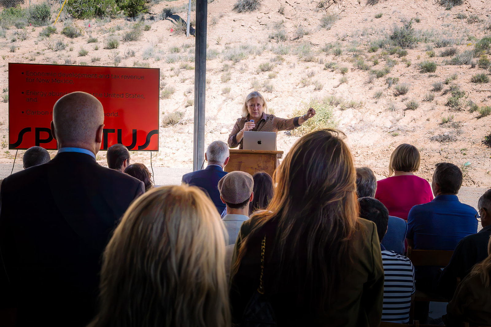 A speaker stands at a podium with a laptop, addressing an audience outdoors, next to a large red presentation board.