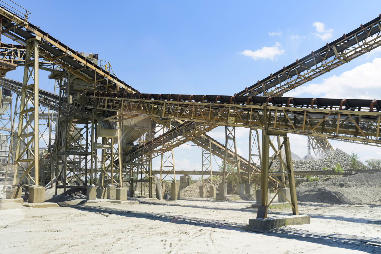 Industrial conveyor belts and metal structures at a gravel processing plant, with piles of gravel and a clear blue sky in the background.