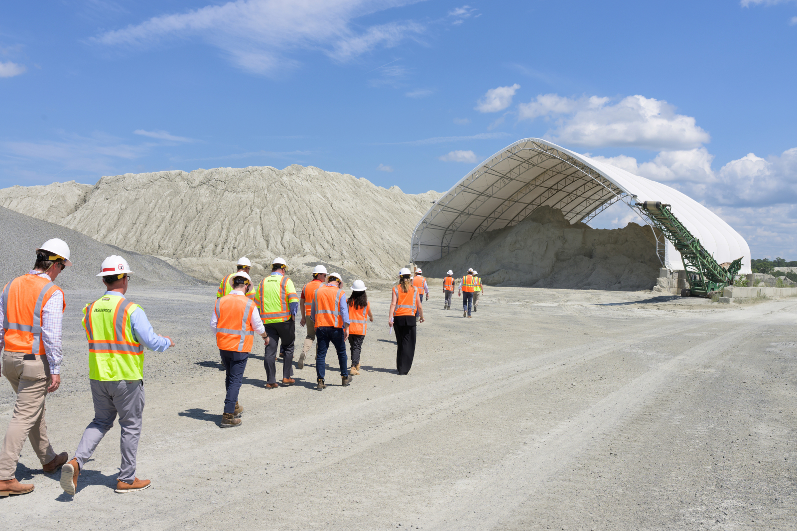 A group of people wearing safety vests and hard hats walking towards a covered industrial facility surrounded by gravel under a clear blue sky.