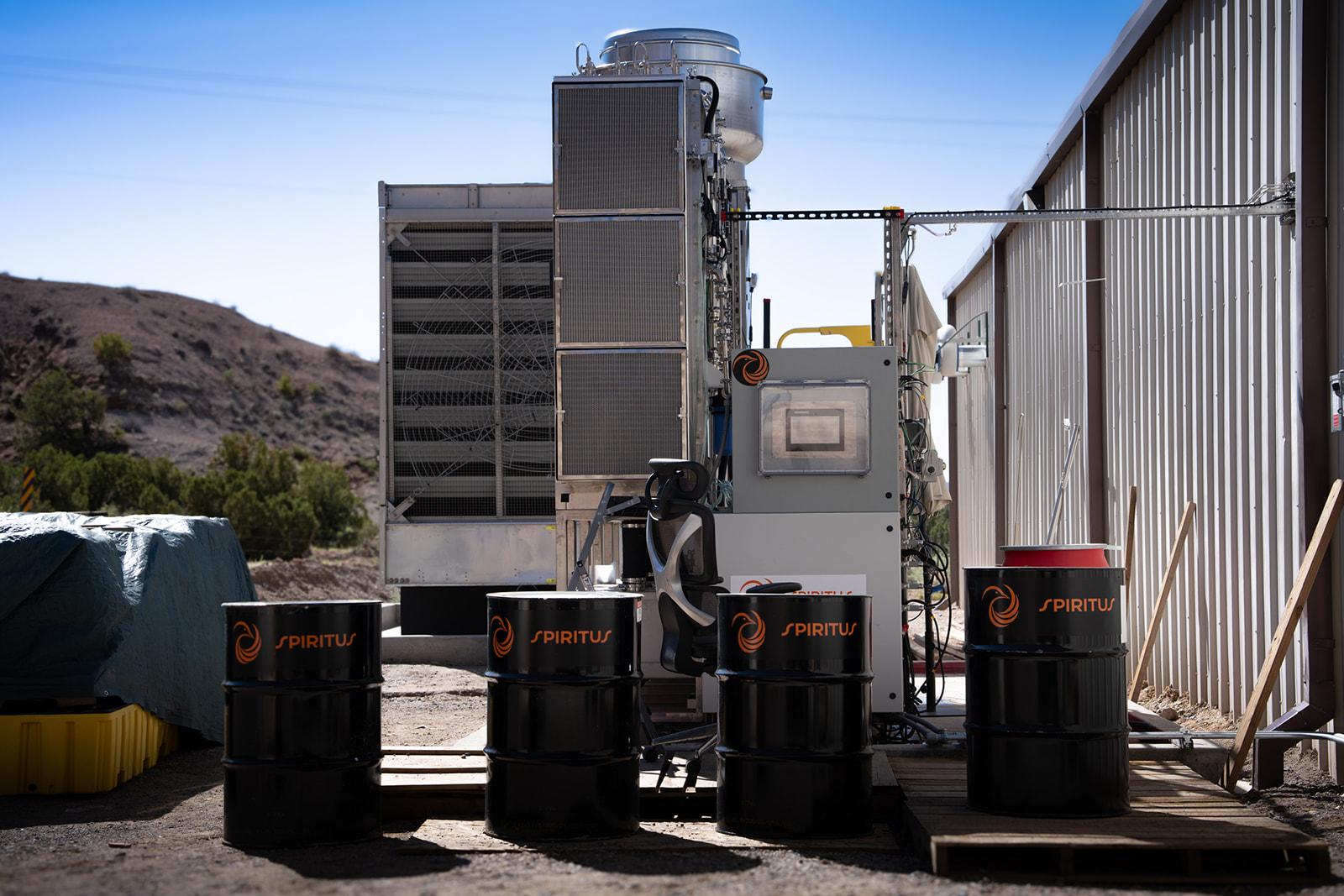 An industrial setup with four black barrels labeled "Spiritus" and a metal equipment structure against a corrugated metal building, set in a desert landscape.