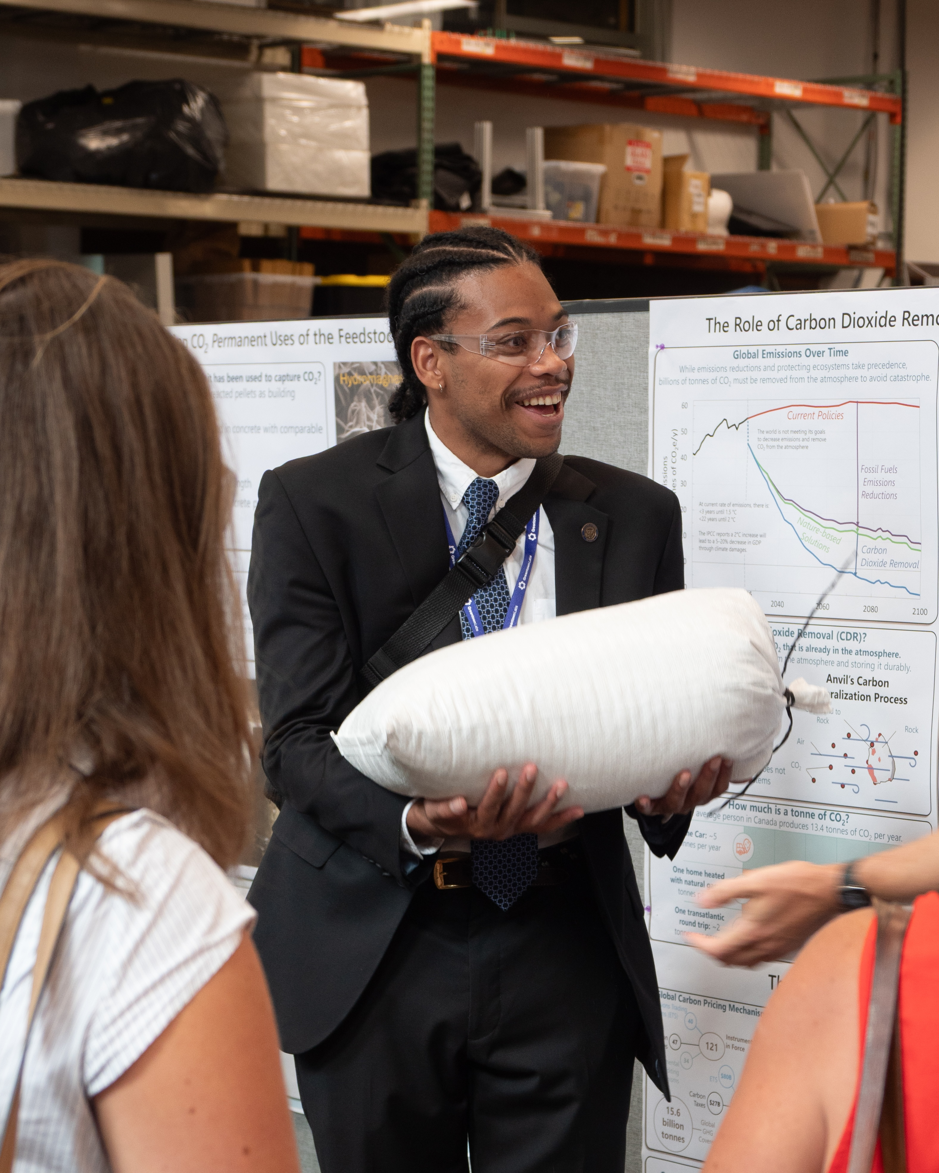A man in a suit and safety glasses holds a cylindrical object, speaking with others in front of charts about carbon dioxide removal.