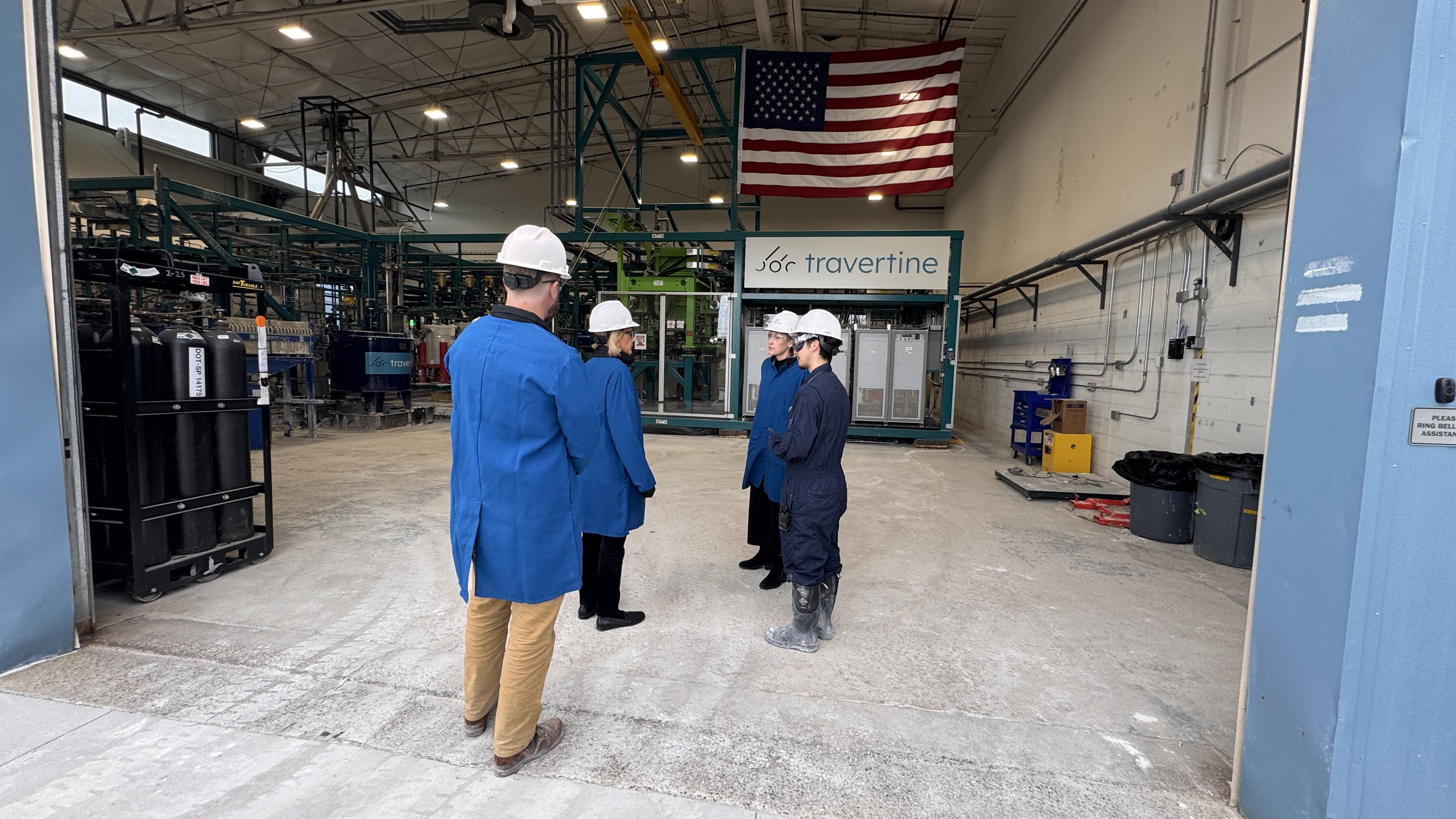 People in blue lab coats and white helmets stand in an industrial facility with a U.S. flag and machinery in the background.