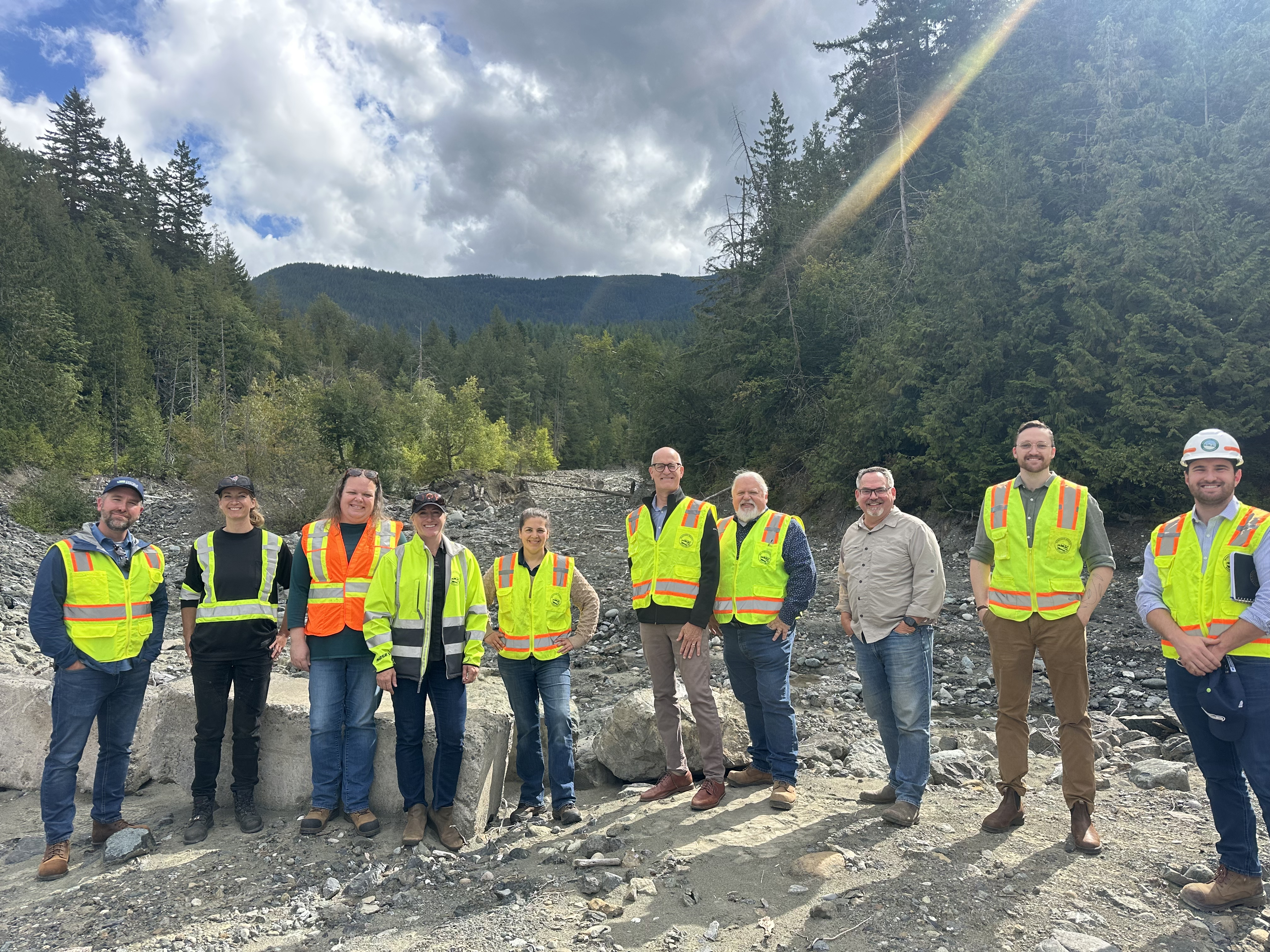 A group of people wearing safety vests and helmets stand on a rocky terrain with trees and mountains in the background.