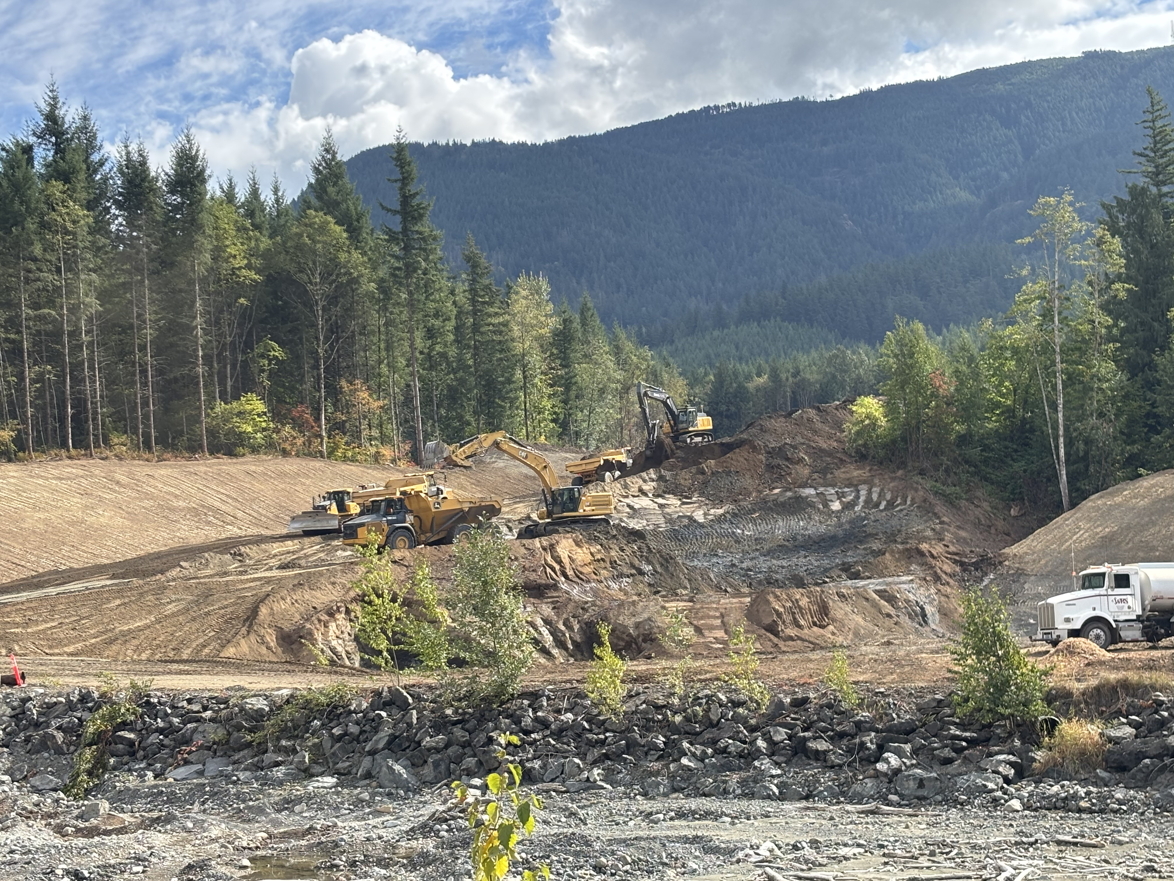 Construction site with excavators and trucks in a forested area, surrounded by mountains under a partly cloudy sky.
