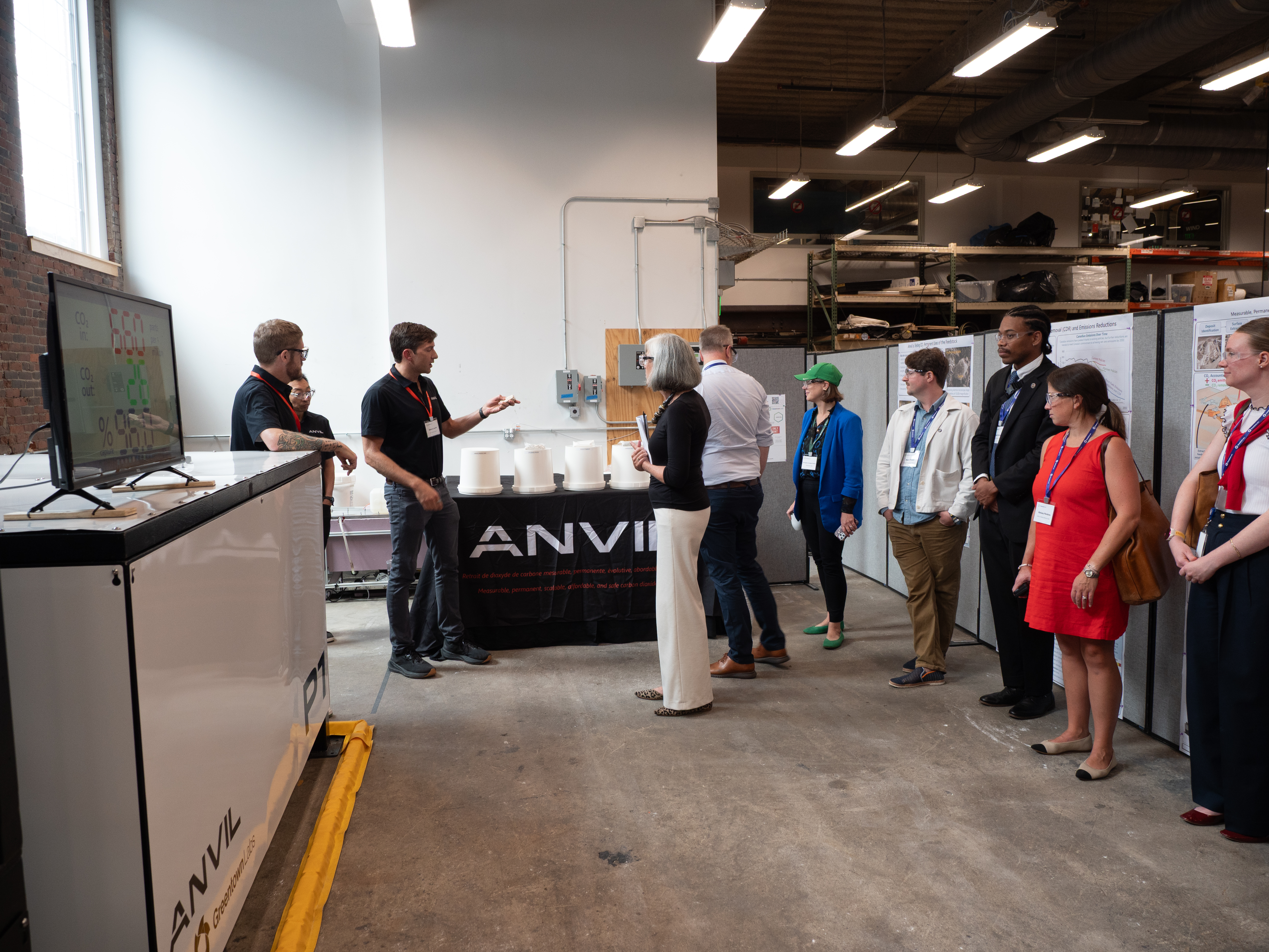 A group of people standing in an industrial space, listening to a presentation by two individuals next to a table and display screen.
