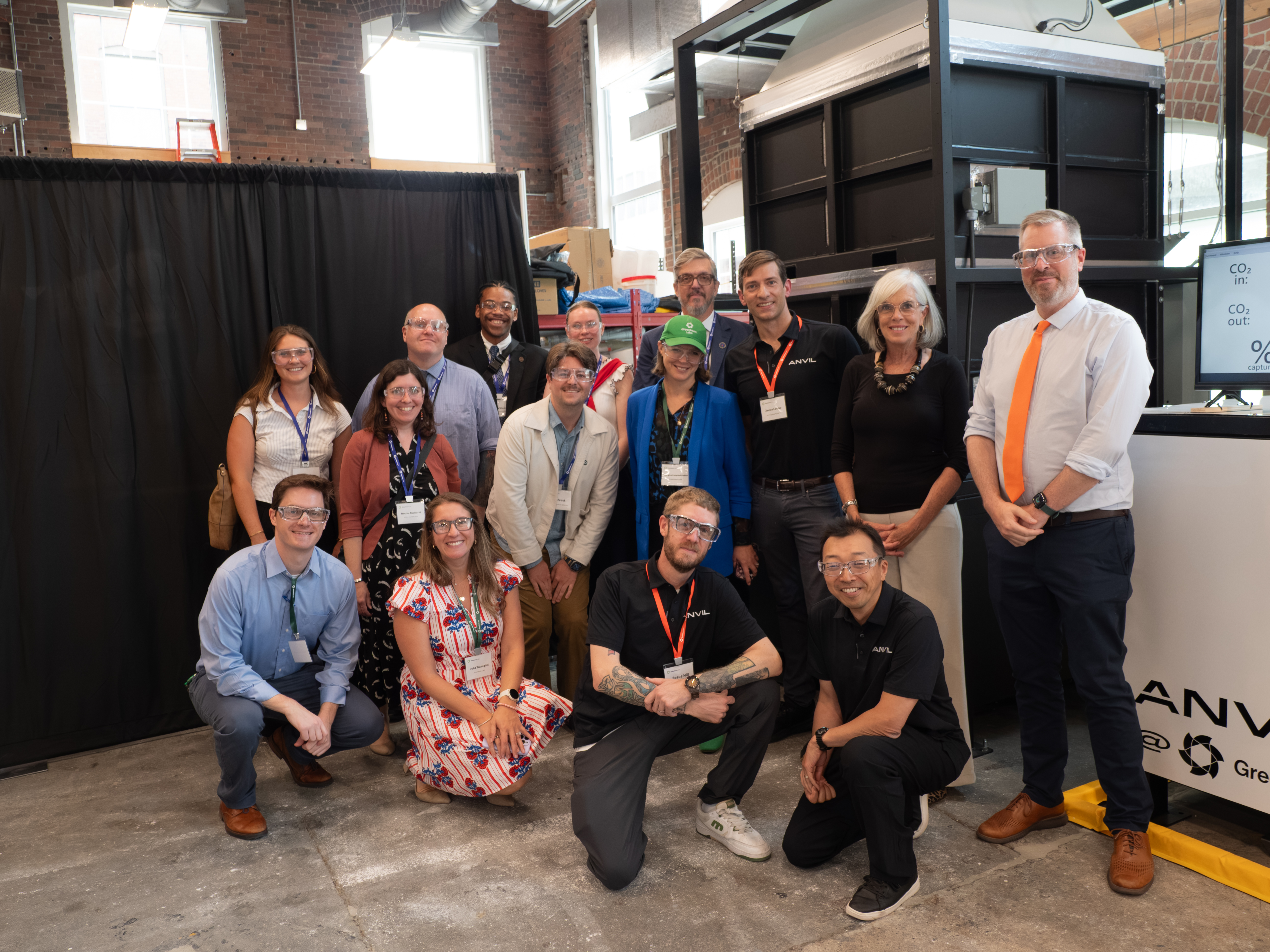 A group of fourteen people pose together indoors in front of industrial equipment, some wearing safety goggles, with a black curtain backdrop.
