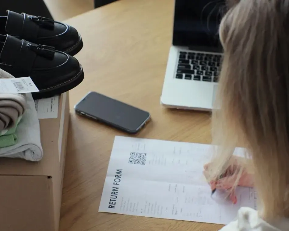 A person fills out a return form at a wooden table, preparing to send back purchased items. A cardboard box holds folded clothes with tags still attached and a pair of black loafers. Nearby are a smartphone, an open laptop, and the partially completed form. The scene reflects a typical home-based product return process in e-commerce.