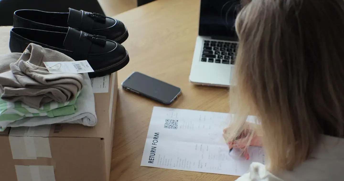 A person fills out a return form at a wooden table, preparing to send back purchased items. A cardboard box holds folded clothes with tags still attached and a pair of black loafers. Nearby are a smartphone, an open laptop, and the partially completed form. The scene reflects a typical home-based product return process in e-commerce.