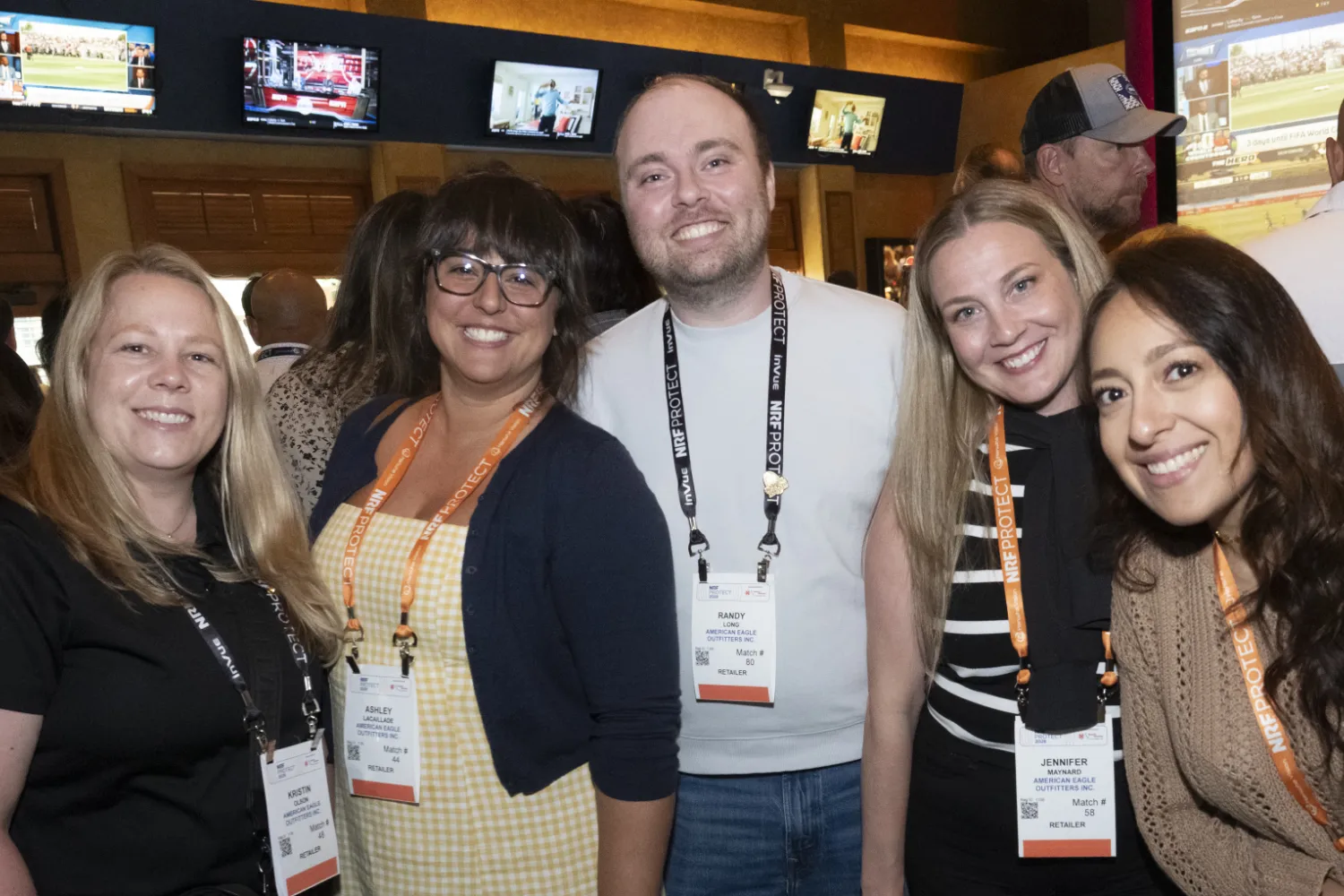 Group of conference attendees wearing NRF PROTECT badges pose together at a networking event with screens in the background.