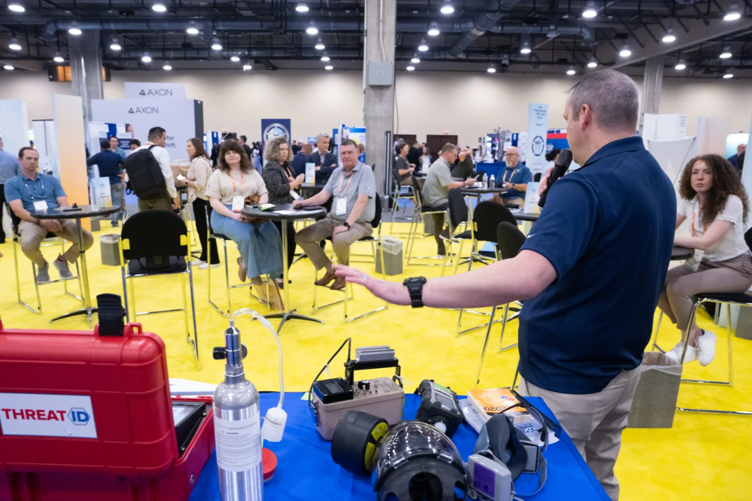 A speaker presents at the NRF Protect Fusion Center, with attendees seated at round tables on a yellow floor, and various equipment displayed on a front table.