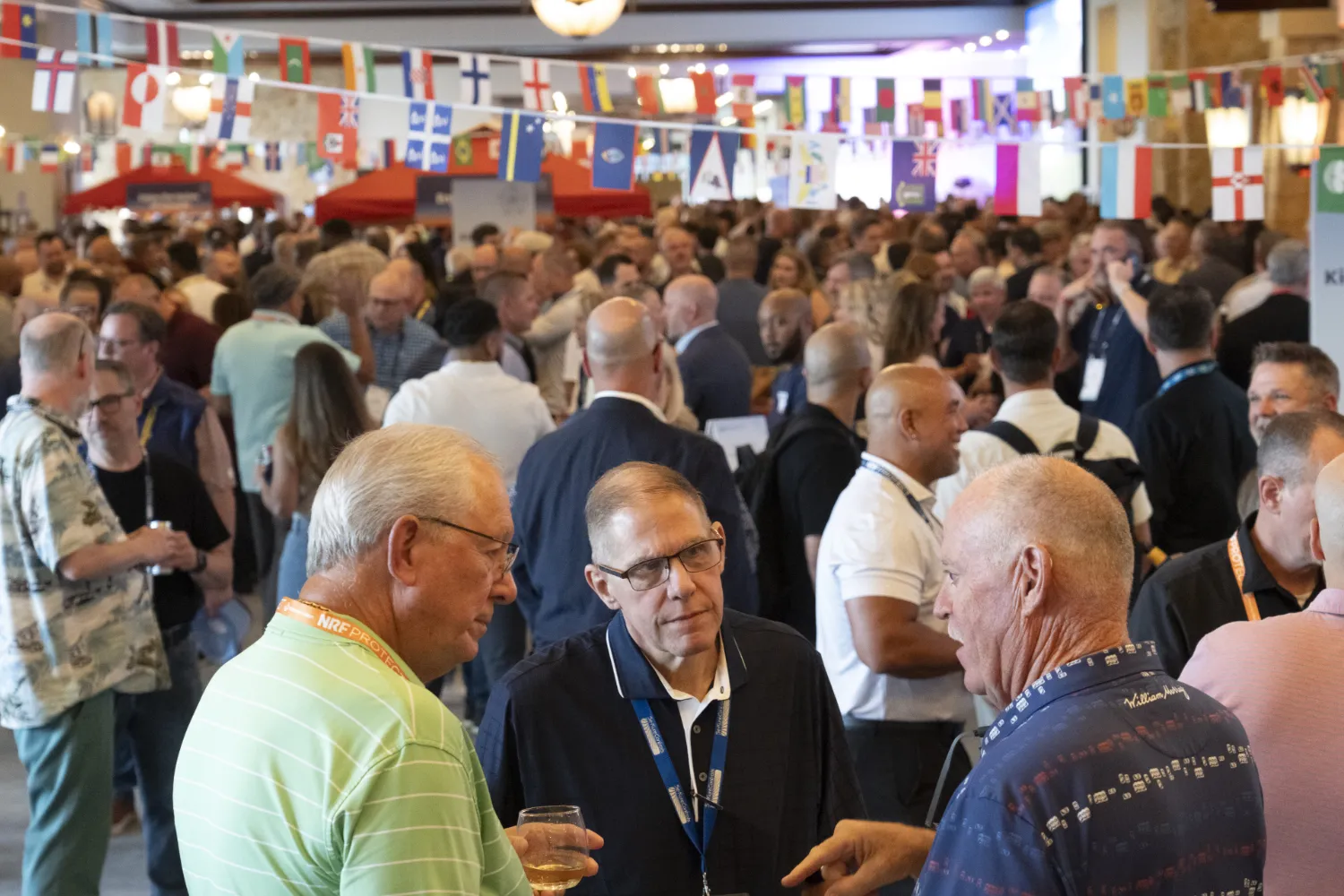 A massive crowd of NRF PROTECT attendees are networking during an evening event. Small, plastic flags from around the world are hanging above their heads, a symbol of the World Cup 2026 theme.