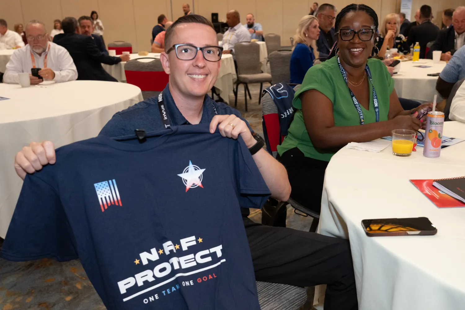 Two NRF PROTECT attendees take a break to hold up a conference t-shirt and smile for the photographer.