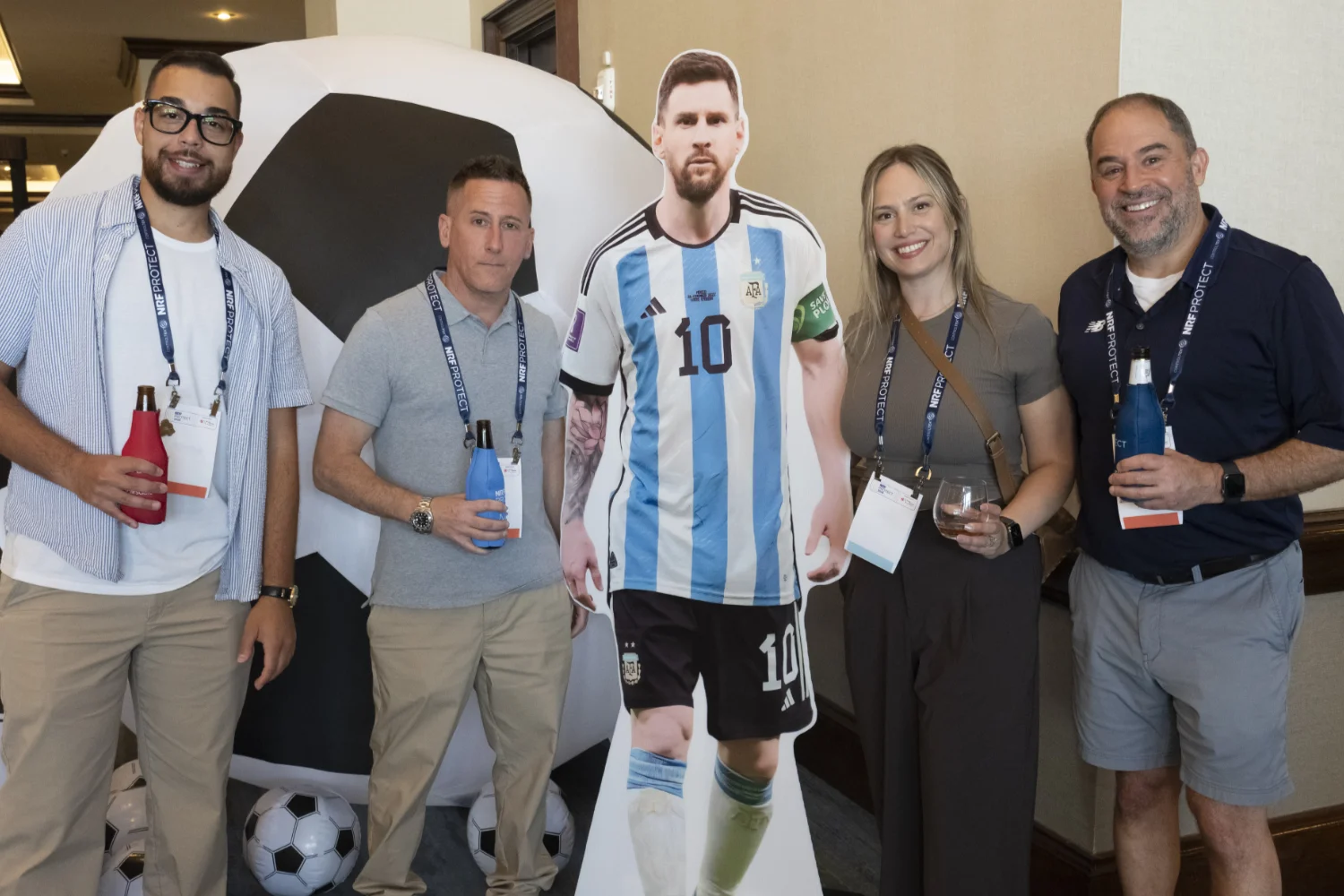 Four people standing around a life-size cardboard cutout of a soccer player Lionel Messi in an Argentina jersey, holding drinks and smiling at NRF PROTECT.