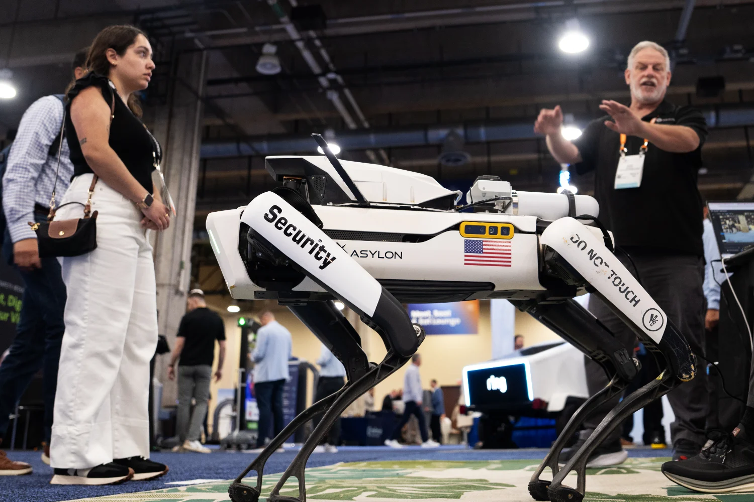 A robotic security dog with "Security" and "Do Not Touch" labels is displayed at the NRF PROTECT Expo, with people observing and a presenter explaining.