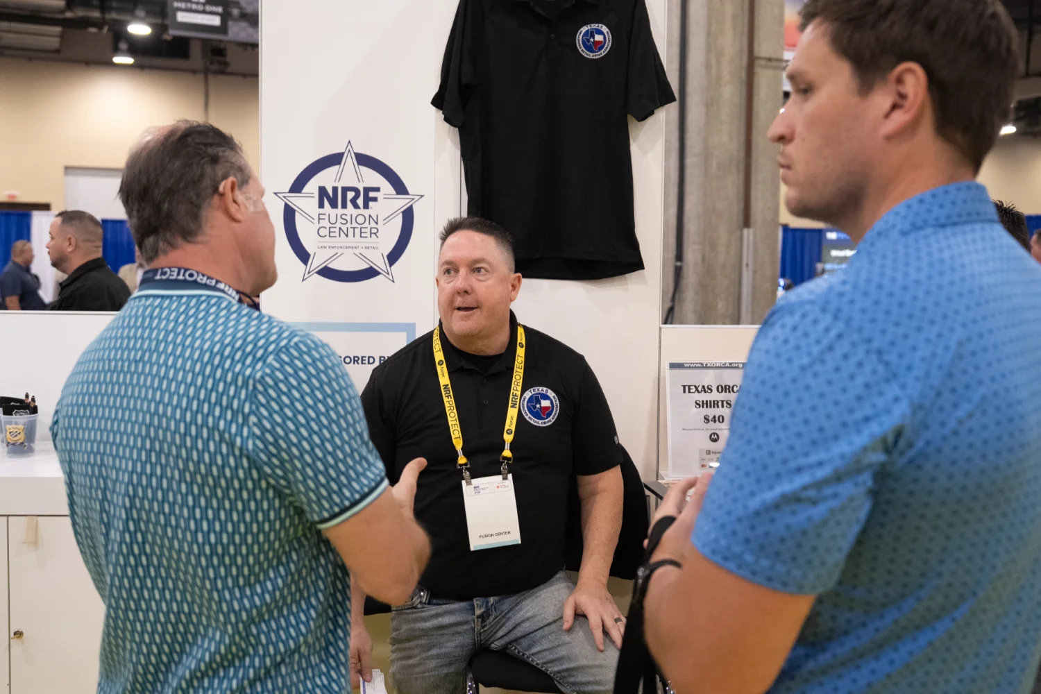 Three men converse at the NRF Fusion Center booth. One is seated, wearing a black shirt with logos, while the others wear blue patterned shirts.