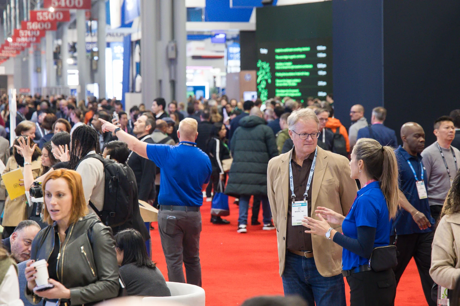 The Big Show expo floor with people walking and conversing and overhead signs indicating booth numbers.