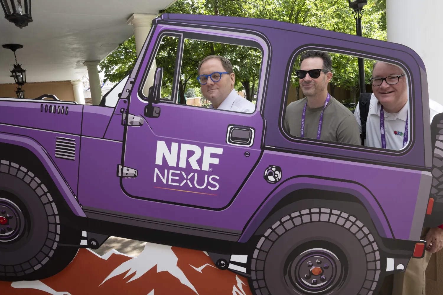 Three men pose for a photo in a cartoon-ish, purple colored standee of a Jeep with the NRF Nexus wordmark written on its side.