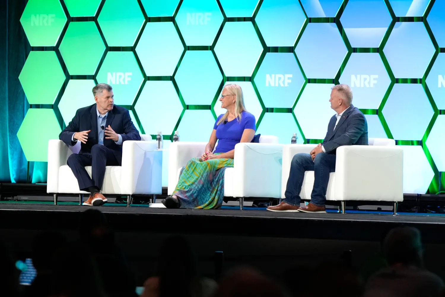 Panel discussion on stage at NRF PROTECT event with three speakers seated in white chairs against a blue and green hexagon backdrop displaying the NRF PROTECT logo.