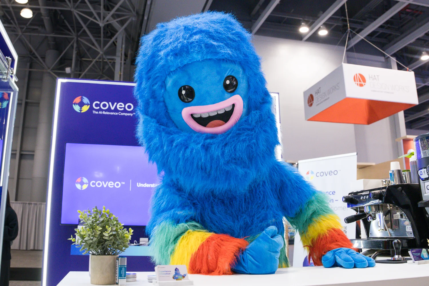 A blue furry mascot with a smiling face and rainbow-colored hands stands at a booth with a small plant and product display.