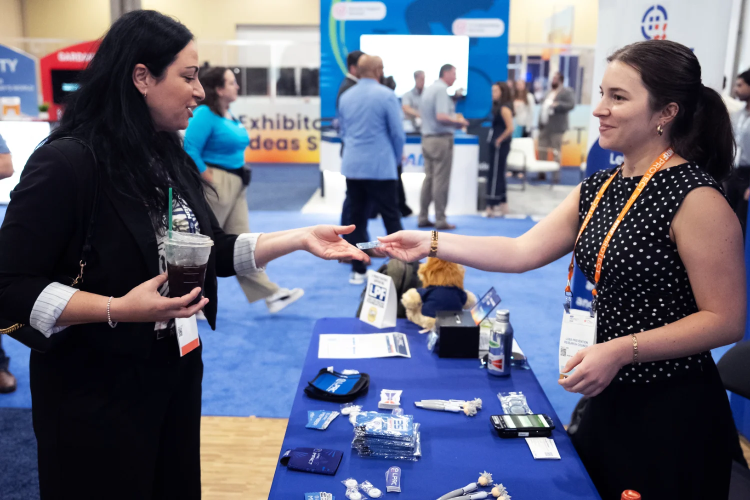 A woman hands another a small giveaway token at a NRF PROTECT EXPO booth. The table between them is loaded with small piles of interesting conference giveaways.
