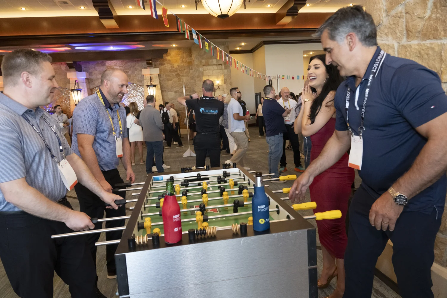 People playing foosball at networking event at NRF PROTECT, with a lively atmosphere and international flags hanging from the ceiling.