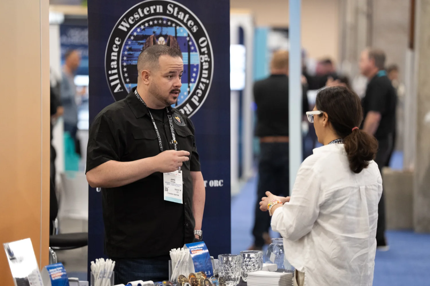 A man in a black shirt talks to a woman in a white blouse at a NRF Protect Fusion Center booth with pamphlets and glassware on the table.