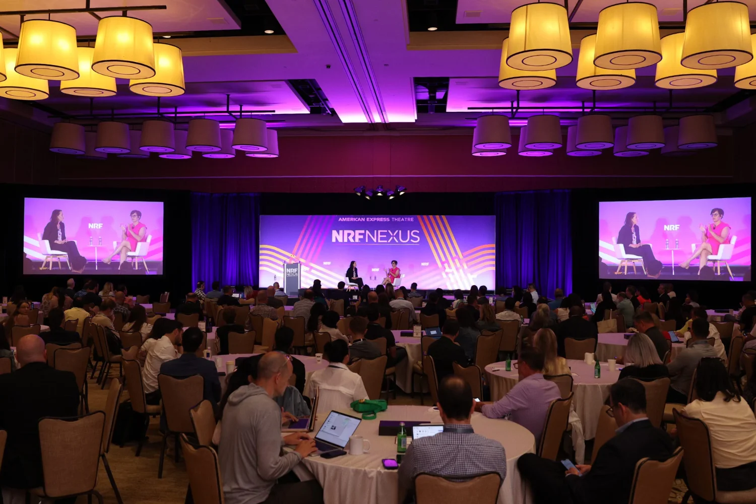 A photograph of the NRF Nexus stage, where two retail experts speak in front of the crowd. The hotel ballroom is darkly lit in purple and pink hues.