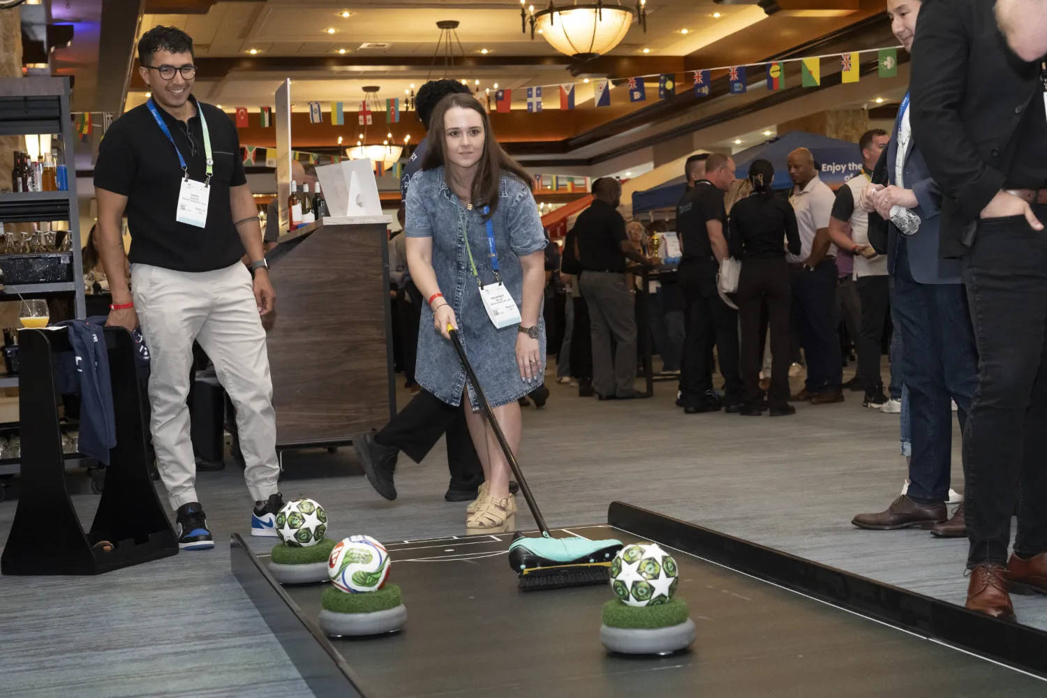 During an NRF PROTECT networking event, a young man and woman use a broom, decorated with a cleat, to play a version of soccer that's akin to shuffleboard. The woman is about to push the ball with the room and is concentrating.