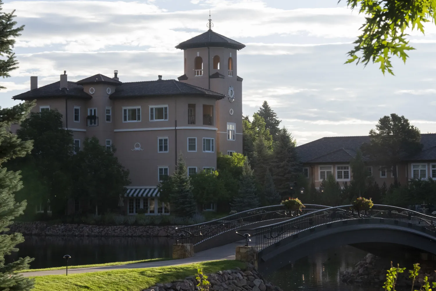 An wide photo of the Broadmoor Hotel. A bridge going over a wide moat is in the foreground. In the background, a clock tower and the hotels accomations.