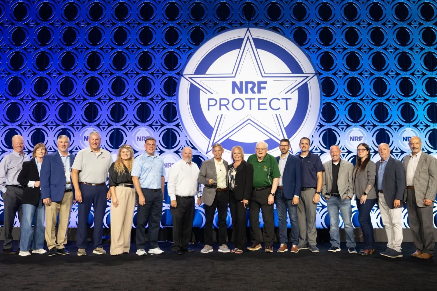A group of 16 professionals in the loss prevention and retail security industry pose with this year's winner of the Ring of Excellence Award at NRF Protect.
