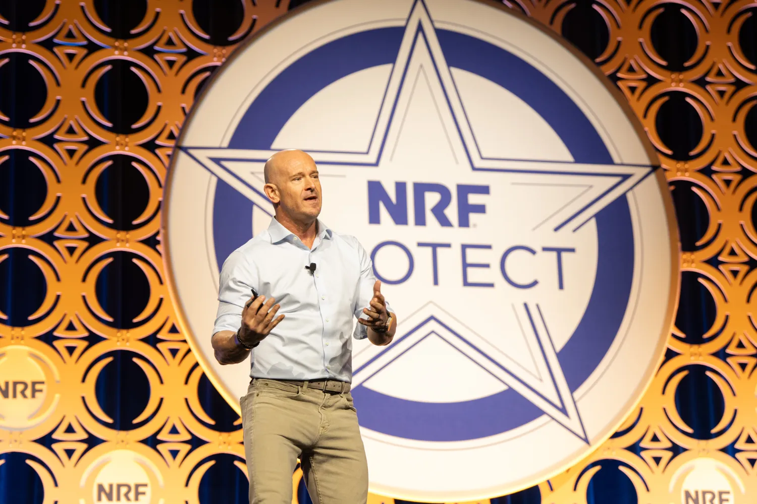 A man emphatically speaks on stage at conference about Loss Prevention. The name of the conference is emblazoned on a sheriff's star on the backdrop behind the man: NRF Protect.