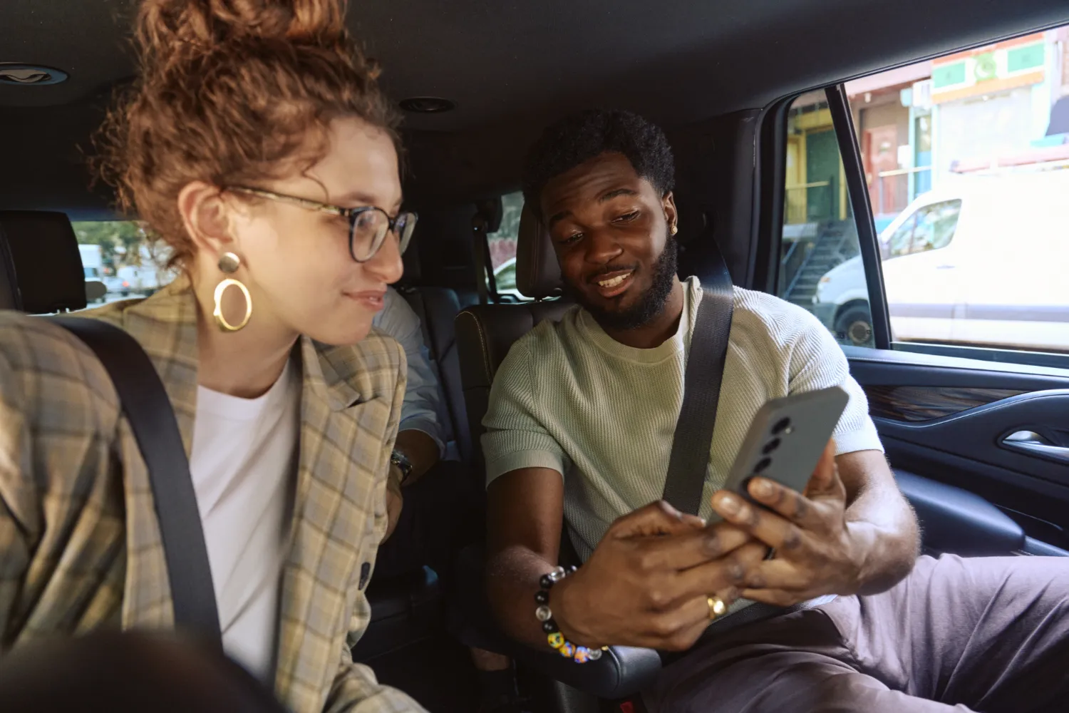 Two people sitting in a Lyft car, smiling and looking at a smartphone. The woman has glasses and a plaid jacket, and the man is wearing a sage-colored shirt.