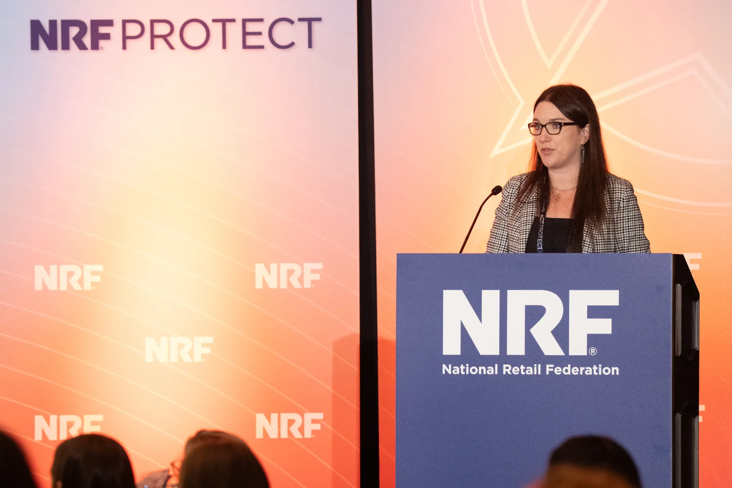 A woman speaks at the NRF Protect podium with "NRF National Retail Federation" branding in front of an audience.