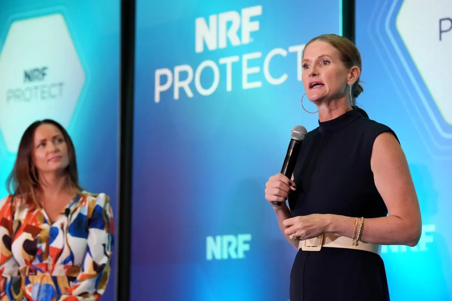 A retail security expert speaks into the microphone to answer a question from the audience. A fellow collegue in the field watches from the left.