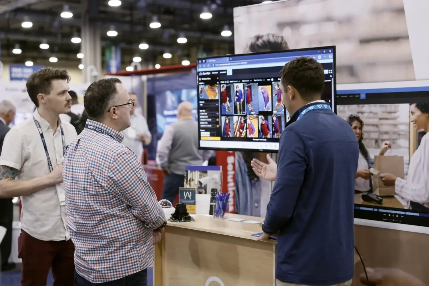 On the NRF PROTECT 2025 Expo floor, we see three men at a booth. One person is demonstrating the latest in cutting edge monitoring technology while two interested people watch listen attentively.