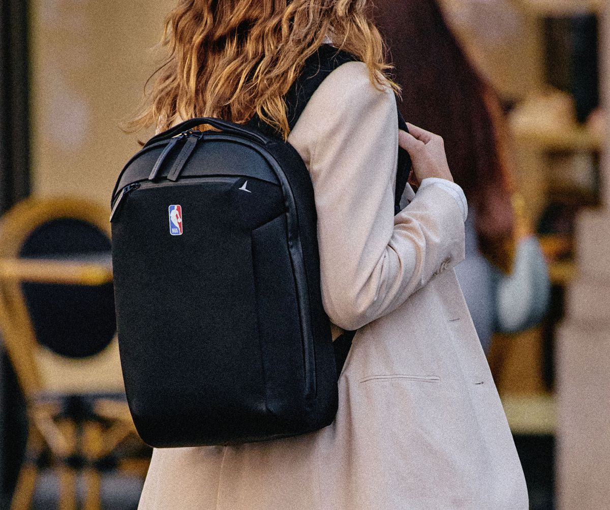 Woman in a cream coat carrying a black Troubadour Apex Backpack embroidered with the NBA logo