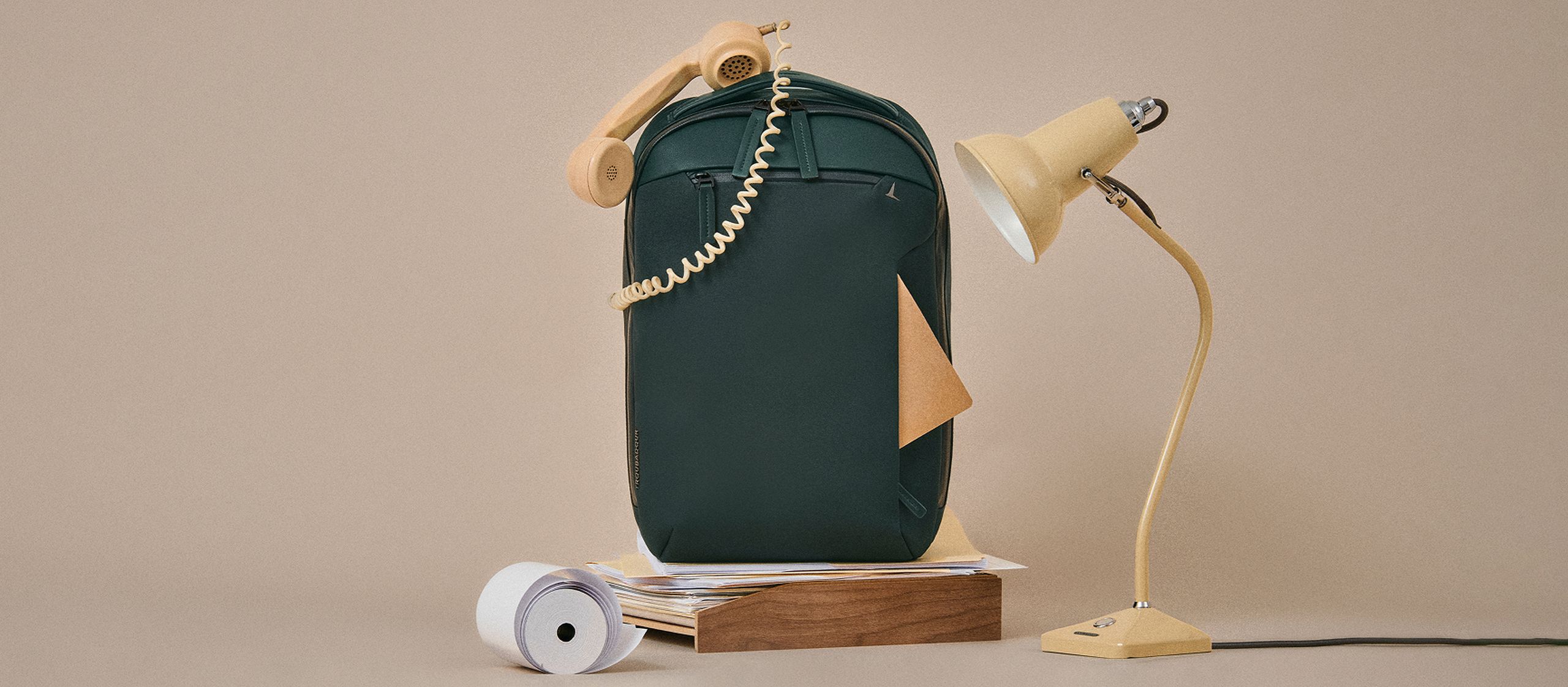 Dark green backpack styled with a vintage beige telephone, a classic desk lamp, paper roll, and wooden block on a neutral background.