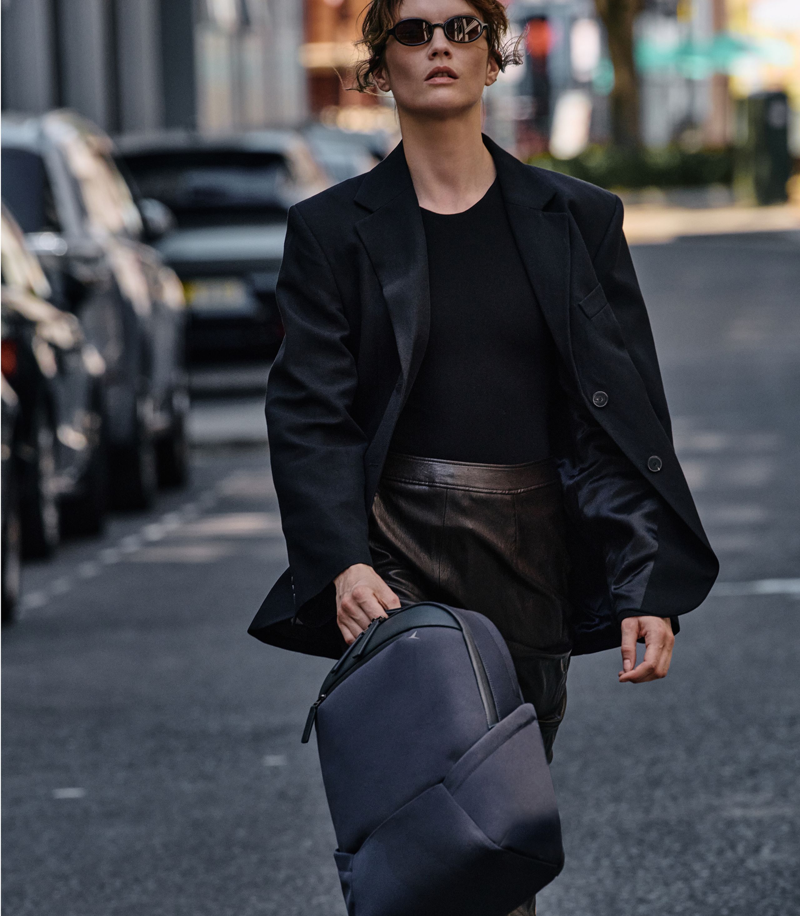 Woman in black blazer and leather skirt walking on city street, carrying a grey structured backpack, wearing small oval sunglasses.