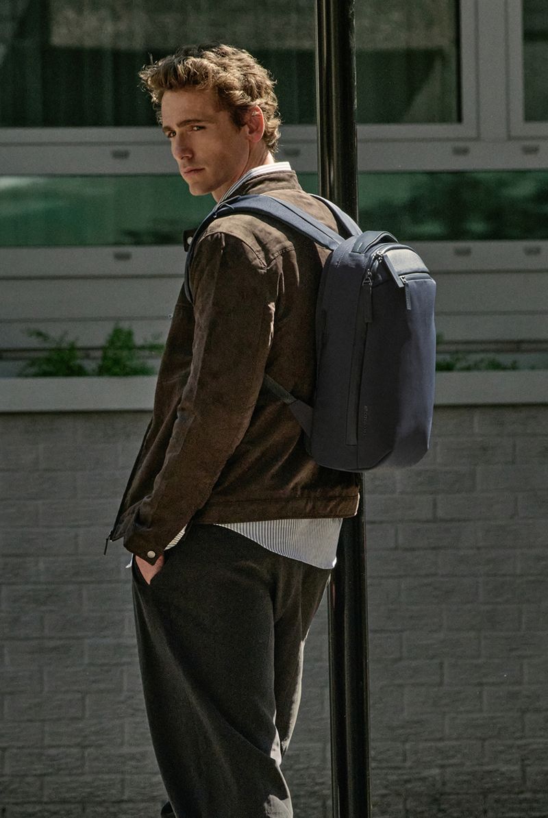 Young man with curly hair wearing a brown jacket and dark pants, carrying a navy blue backpack, leaning against a pole outdoors.