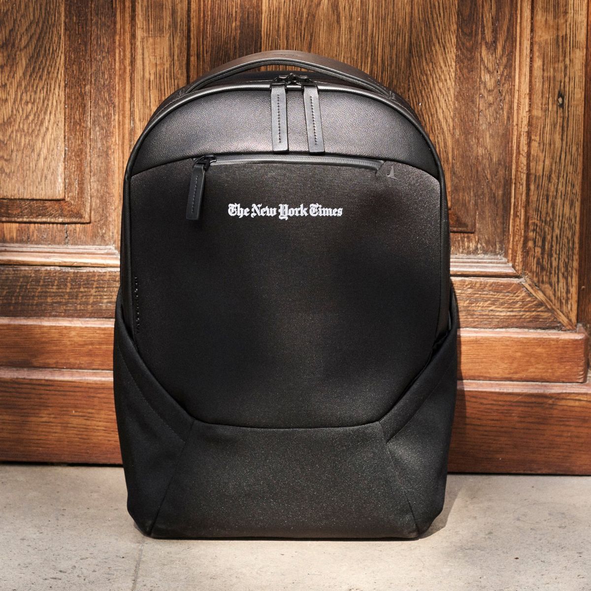 Troubadour Apex Backpack 4.0 with The New York Times logo embroidered on the front, standing against an oak door
