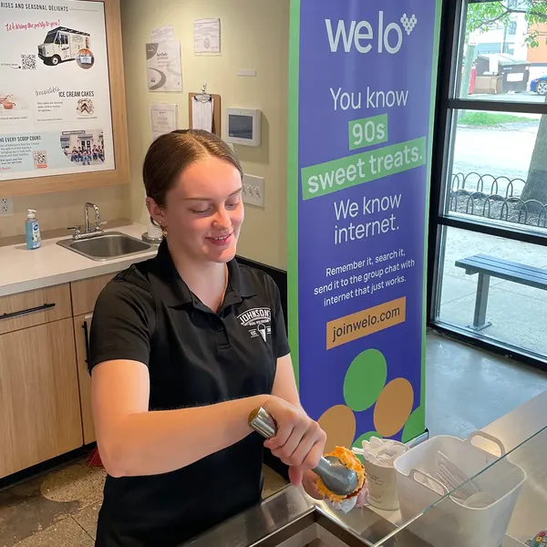 An image of an employee at Johnson’s Ice Cream scooping orange ice cream into a cone next to a purple welo banner.