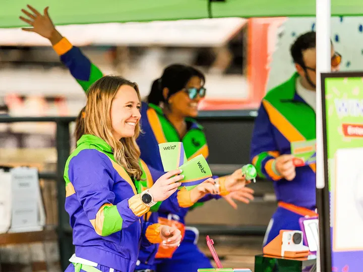 An image of 3 welo squad members in 90s-style blue, green, and orange tracksuits, smiling and handing out green branded cards.
